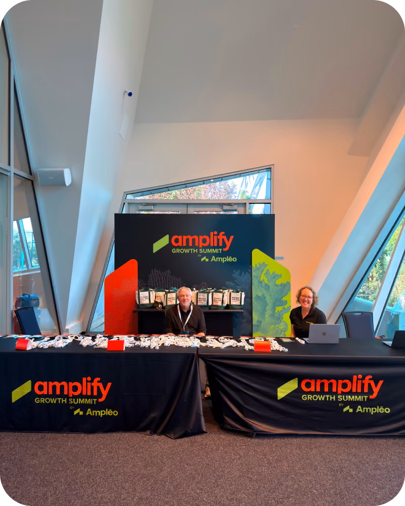 Two smiling people seated behind black tables covered with lanyards and event materials at the Amplify Growth Summit by Ampléo registration or information desk in a modern, bright venue with large angled windows.