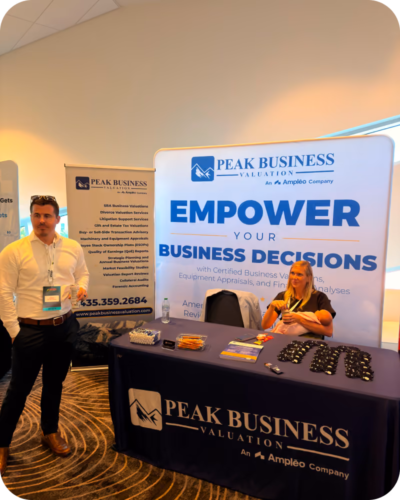 Trade show booth for Peak Business Valuation with a man standing on the left holding a lanyard and woman sitting behind a table on the right holding a baby, promotional items on the table, and banners displaying business valuation services.