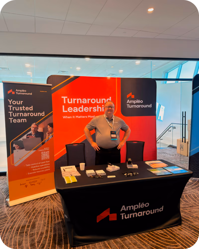 Man standing behind a black table with promotional materials at an Ampleo Turnaround booth featuring banners that say 'Turnaround Leadership When It Matters Most' and 'Your Trusted Turnaround Team', in a modern indoor setting.