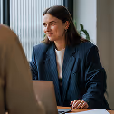 Woman wearing a dark blazer smiles and looks to the side while sitting at a desk.