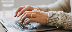 Close-up of hands typing on a laptop keyboard with a gray sweater sleeve visible.