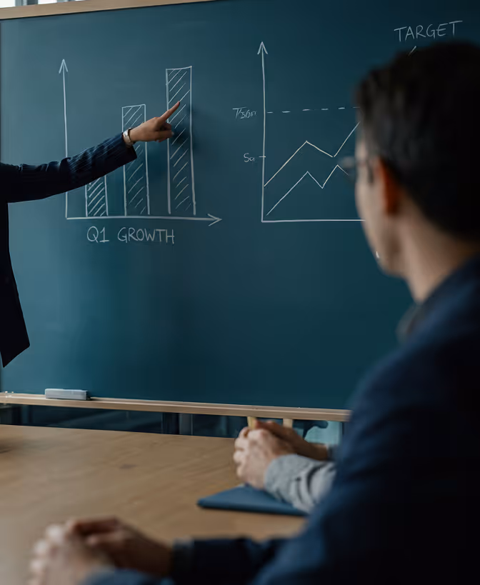 Person pointing at bar chart on chalkboard showing Q1 growth while another person watches.