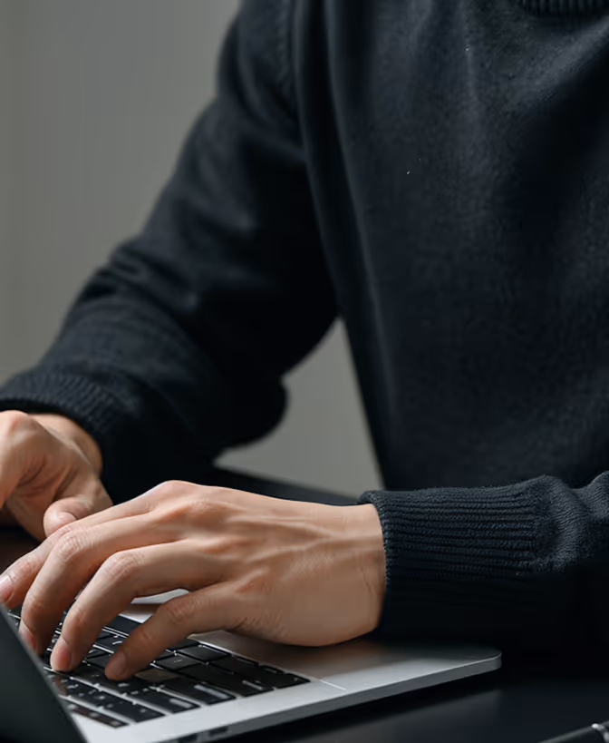 Person wearing black sweater typing on a laptop keyboard at a dark desk.