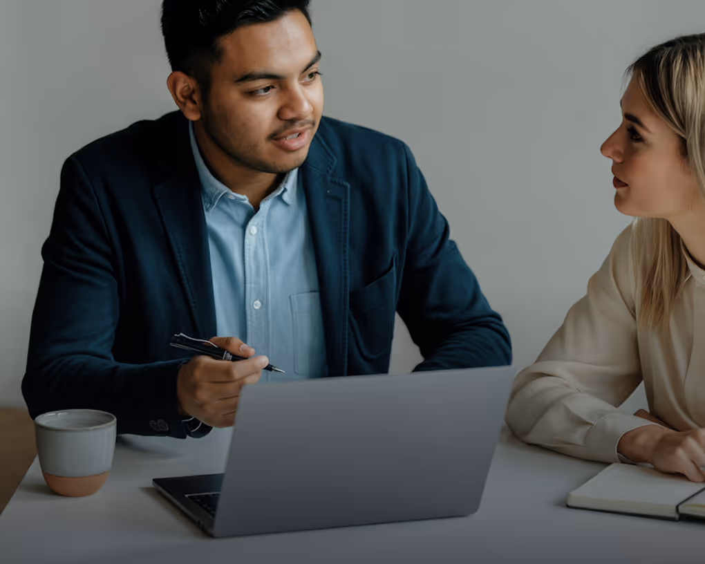 Two coworkers discussing work at a laptop in a modern office setting.