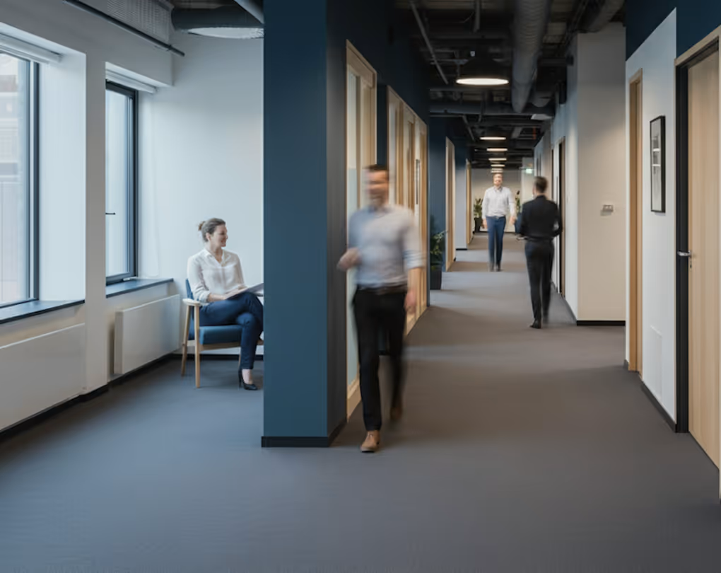 Woman sitting by window and three blurred people walking in a modern office hallway.
