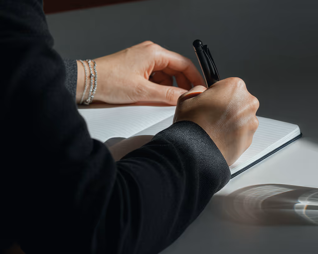 Person wearing a black sleeve writing with a pen on a blank notebook at a white desk.