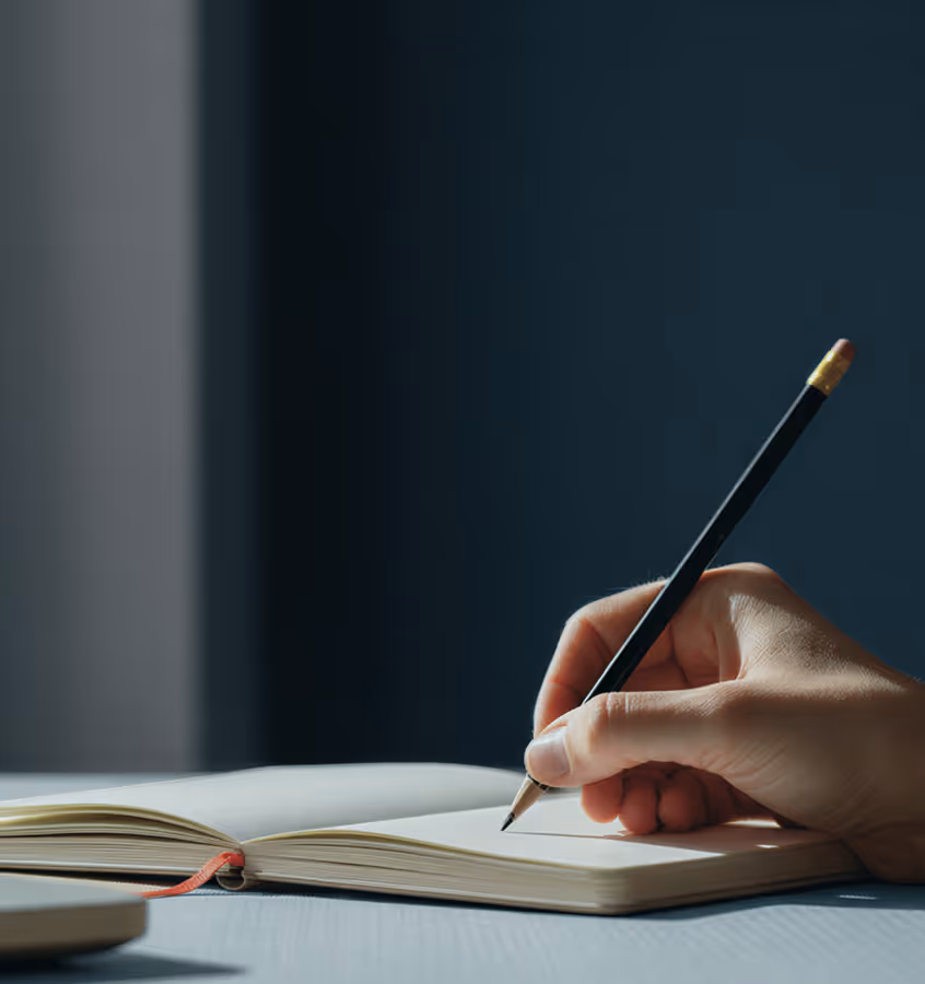 Hand holding pencil poised to write on an open notebook on a table with soft lighting.