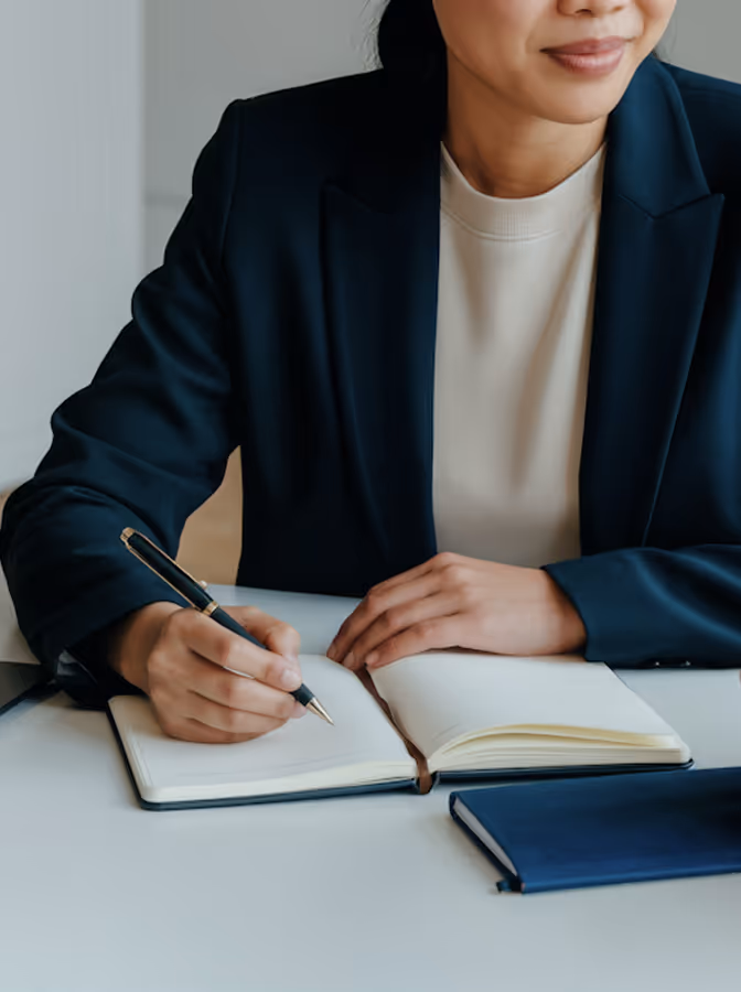 Person in dark blazer writing in an open notebook on a white table with another closed book nearby.