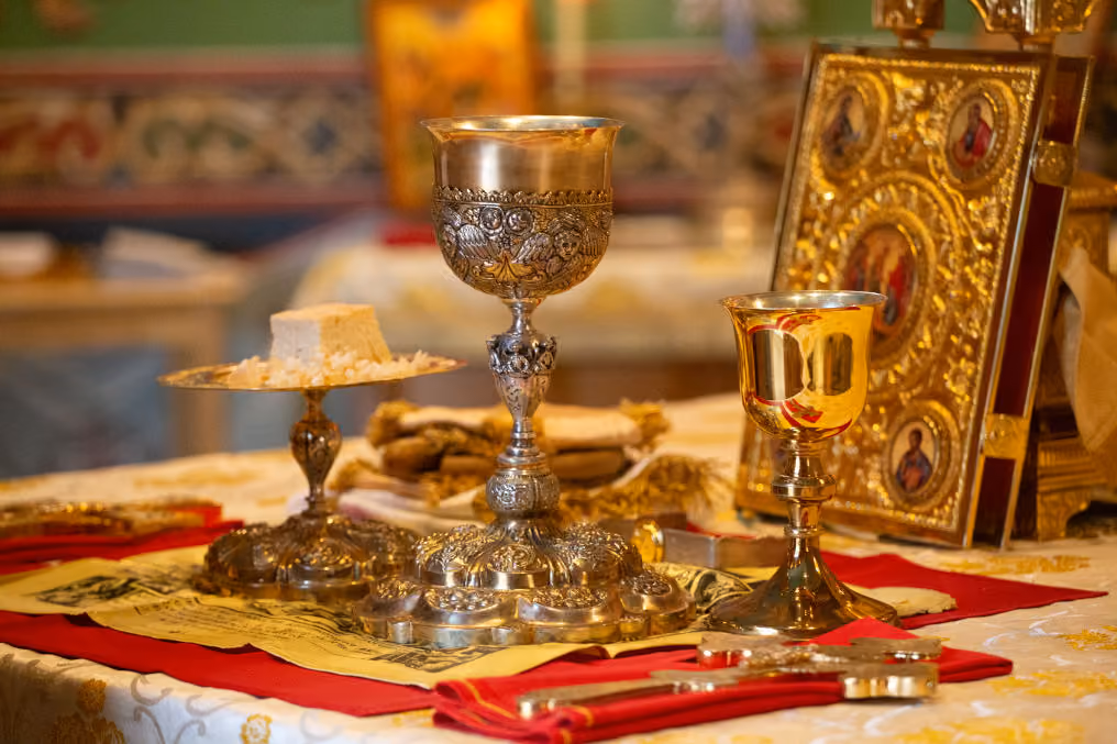 Ornate religious chalices and plate with bread arranged on a decorated altar cloth, with a gold-covered book featuring religious icons in the background.