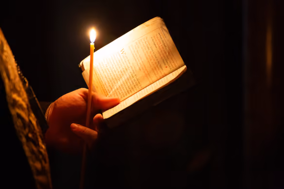 Person holding a lit candle and an open book, illuminated by the candlelight in a dark setting.