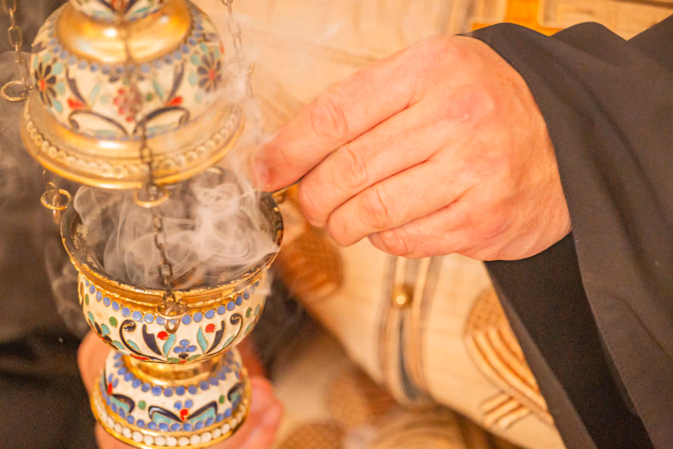 Close-up of a person holding a decorated brass incense burner with smoke rising from it, wearing black sleeves and a garment with gold embroidery.