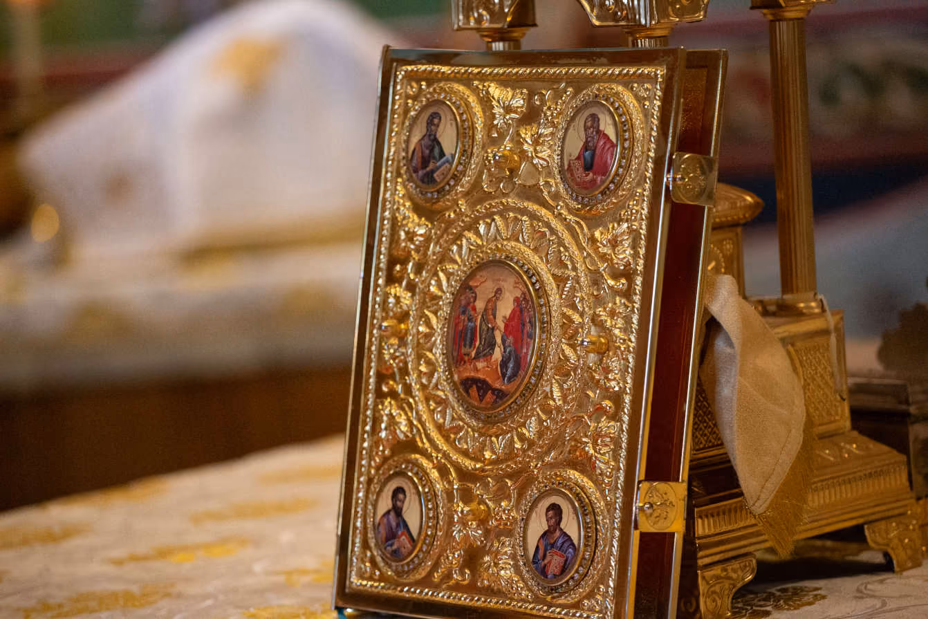 Ornate gold-covered religious book or icon with four smaller painted portraits of saints in each corner and a central scene depicting biblical figures, placed on a decorated surface in a church setting.