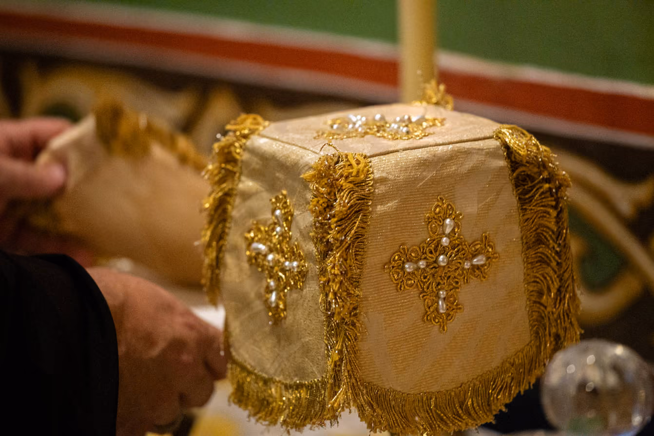Close-up of a gold embroidered cloth with ornate crosses decorated with pearls and gold fringe, placed on a religious altar with a candle visible behind it and hands partially seen.