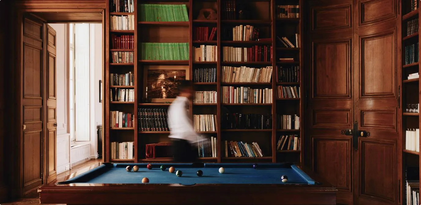 Une salle élégante avec une grande bibliothèque en bois remplie de livres, une table de billard au centre avec des boules disposées dessus et une personne floue en mouvement près de la table.