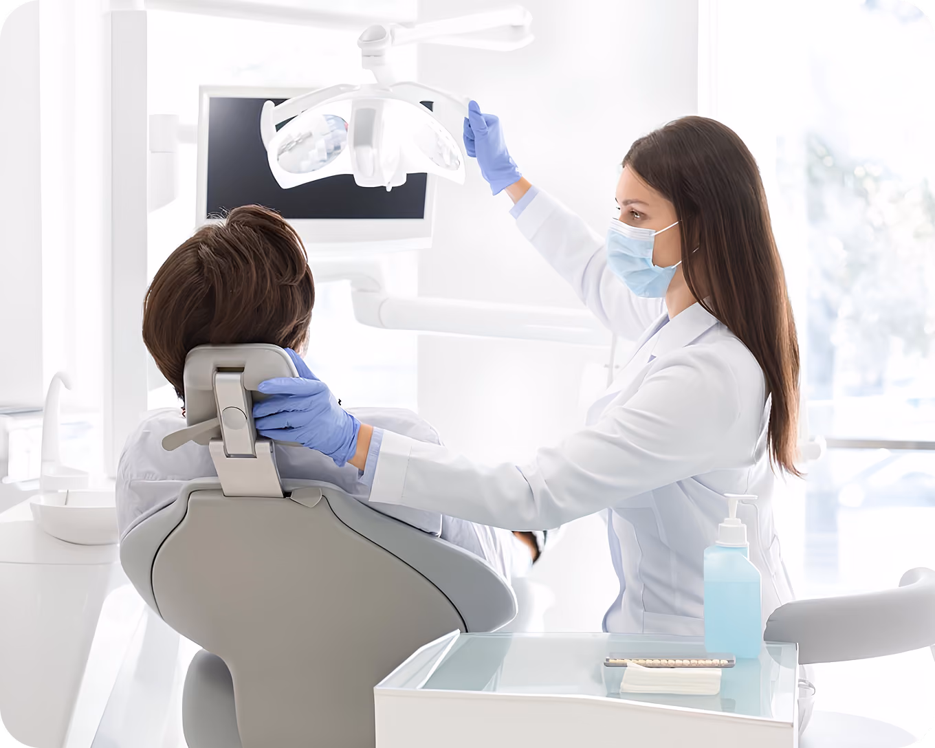 Dentist wearing mask and gloves adjusting dental light over patient sitting in chair.