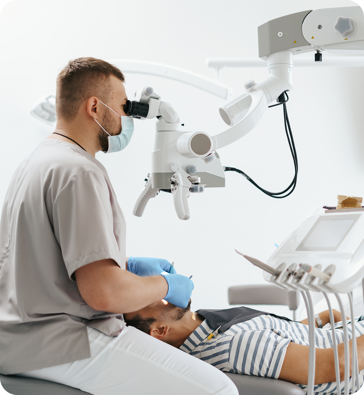 Dentist wearing mask and gloves using a microscope to examine a patient's mouth in a dental clinic.
