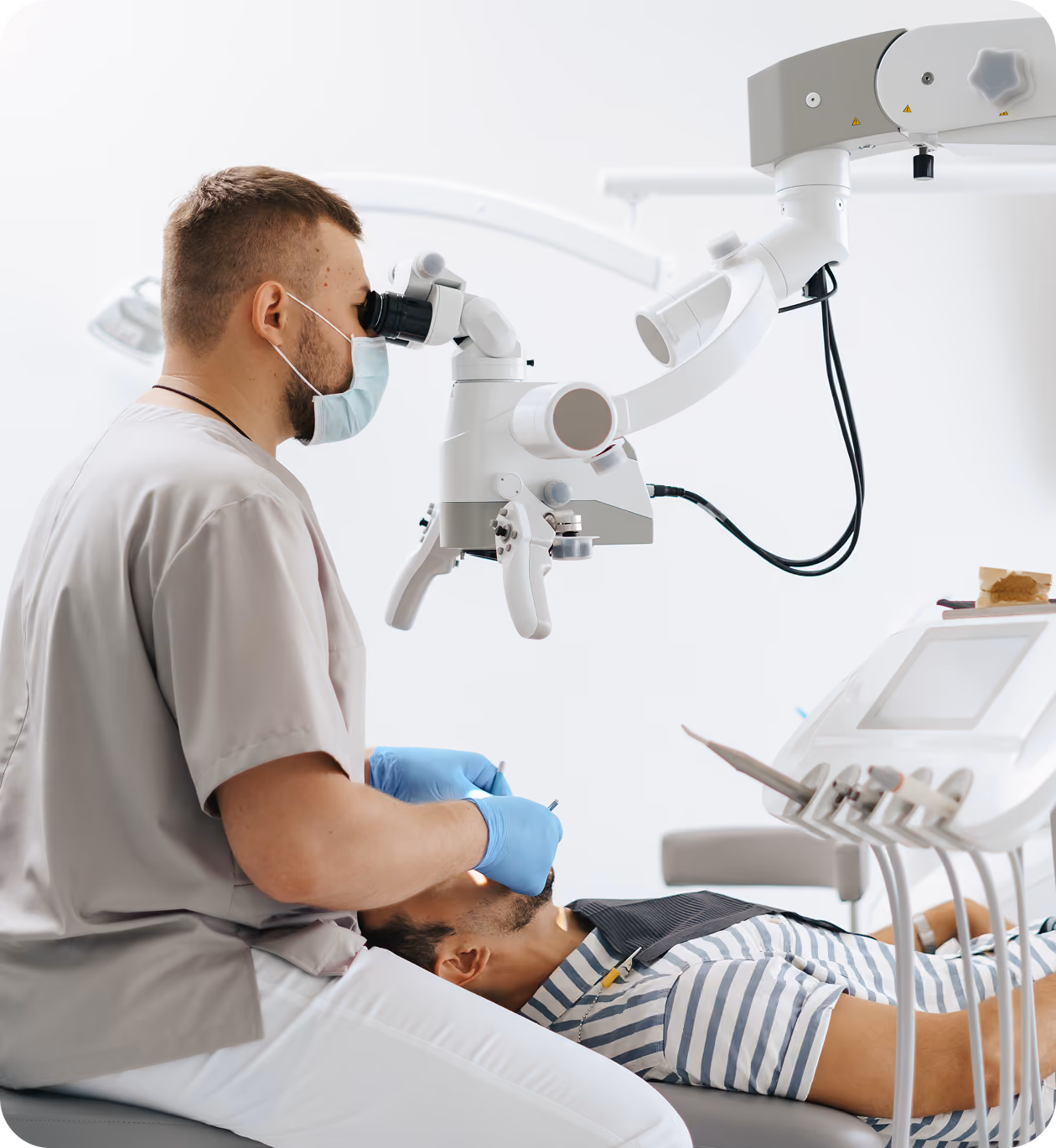 Dentist wearing mask and gloves using a microscope to examine a patient's mouth in a dental clinic.