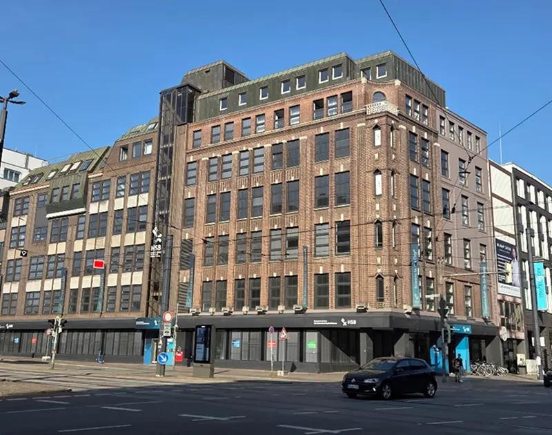 Großes Backsteingebäude an einer Kreuzung bei sonnigem Himmel mit Auto und Fahrradfahrern.