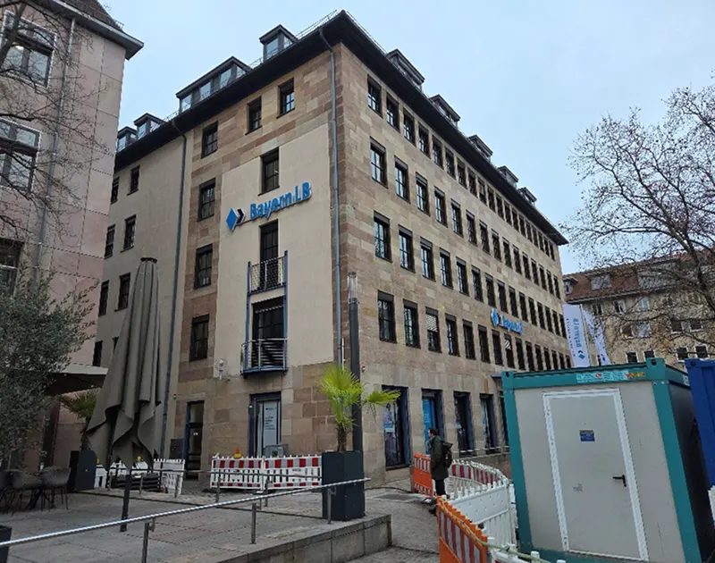Gebäude mit BayernLB-Schriftzug und Fenstern an grauem Himmel, Bäume und Bauzäune im Vordergrund.