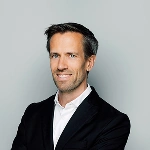 Porträt eines lächelnden Mannes (Andreas Quaas) mit dunklem Jackett und weißem Hemd vor grauem Hintergrund.