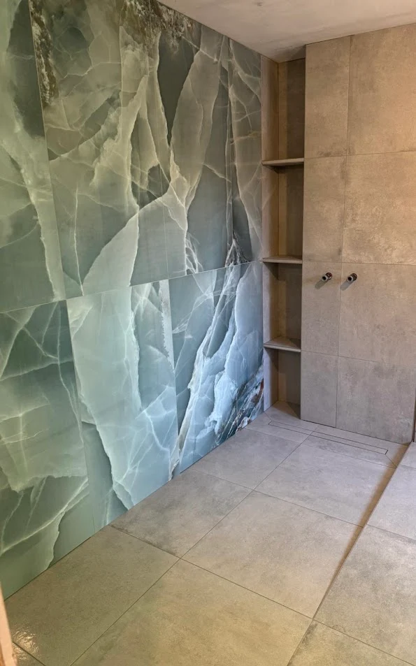 Corner of a tiled room with large green and white marble wall, beige floor tiles, and built-in shelves.