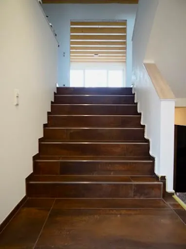 Indoor brown tiled staircase with white walls leading to a bright window and wooden ceiling.