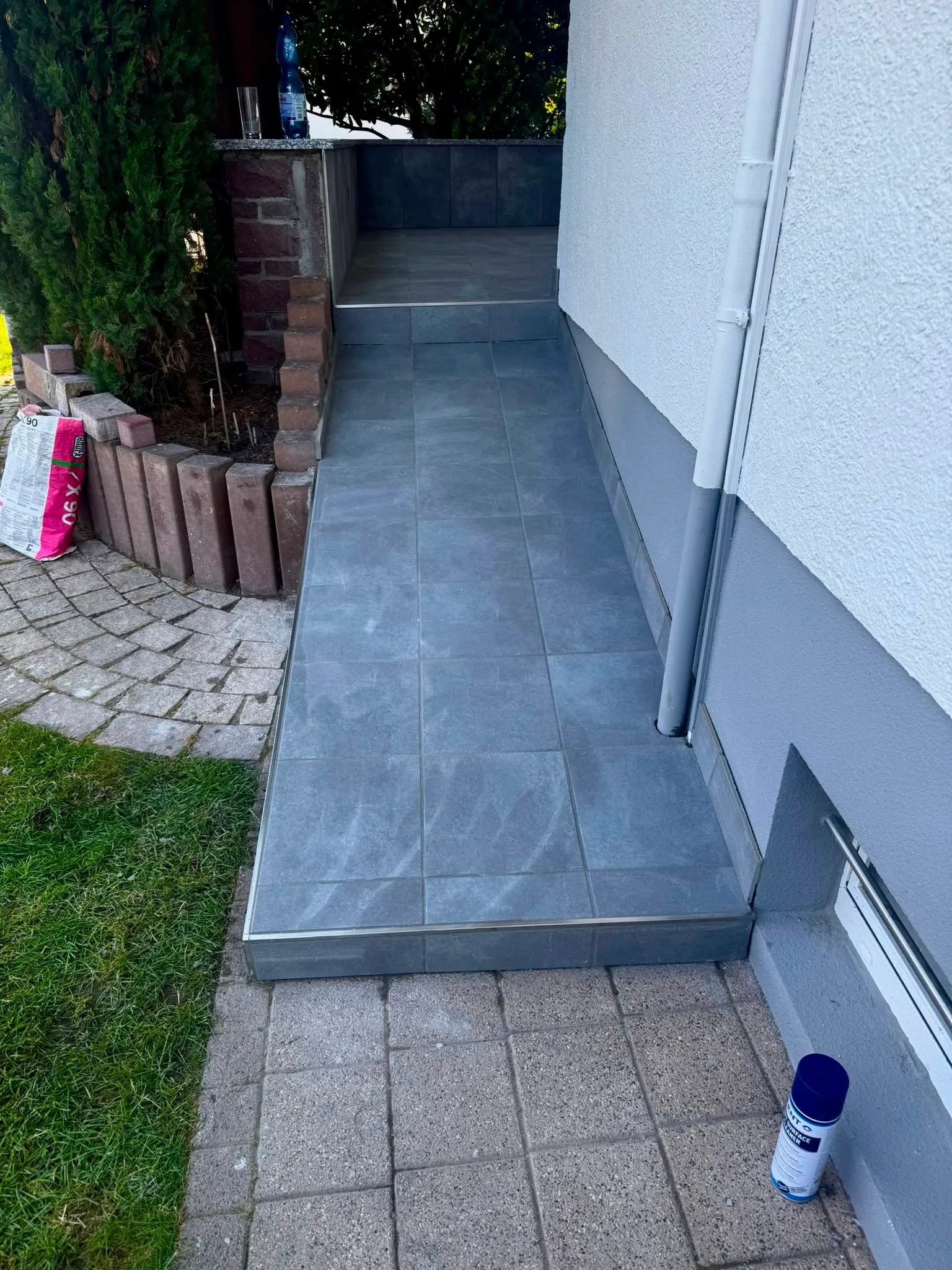 Grey tiled ramp and steps next to a white and grey house wall with plants and grass nearby.