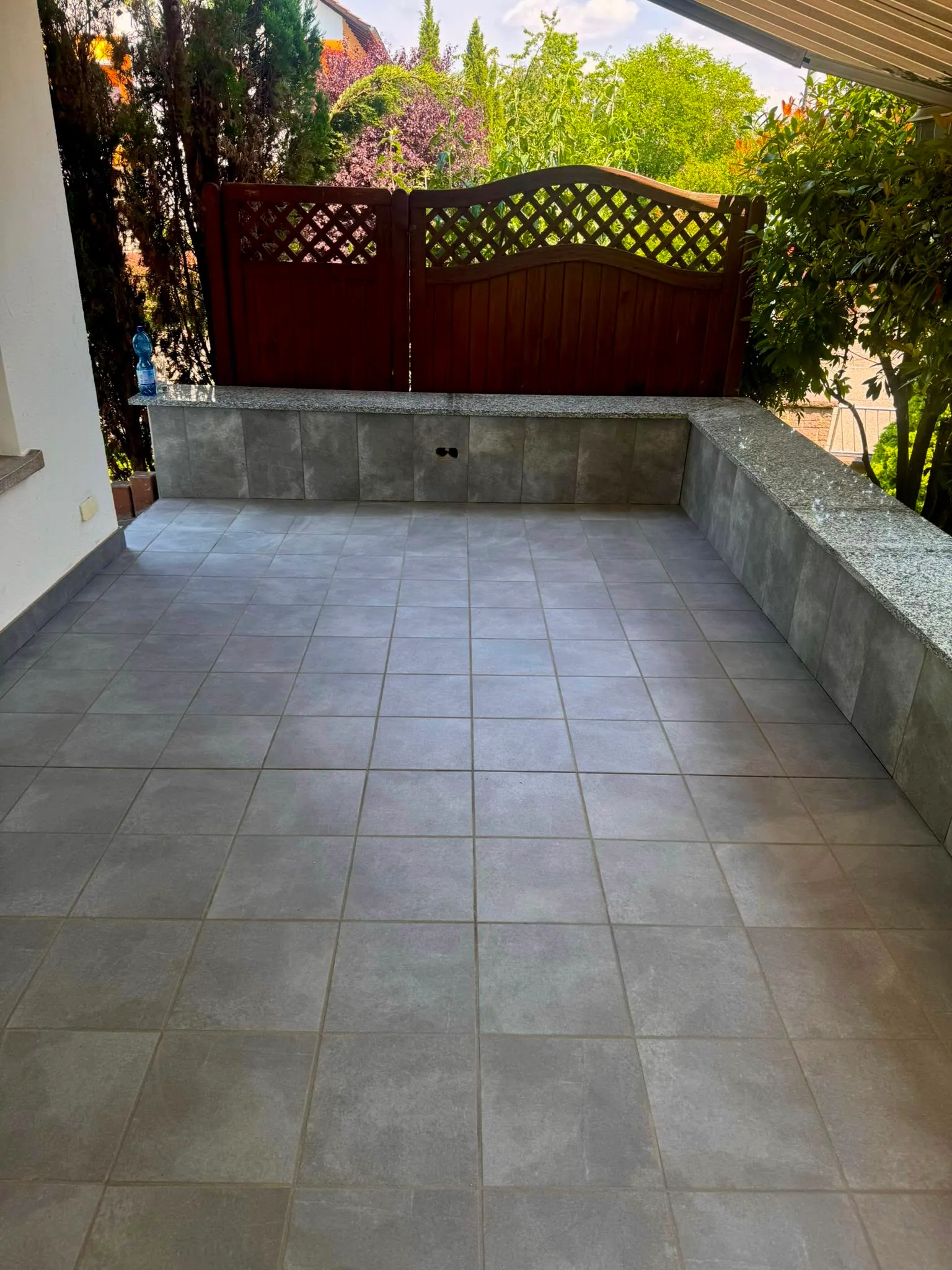 Empty outdoor tiled patio with built-in bench and wooden privacy fence surrounded by greenery.