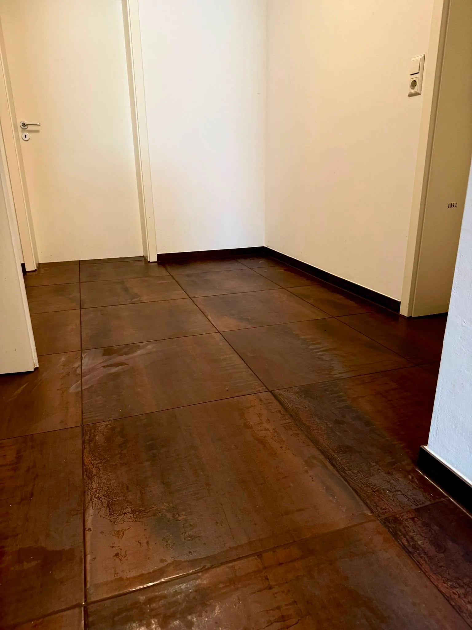 Indoor corner with white walls, closed white door, and brown textured tiled floor.