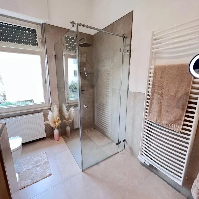 Modern bathroom with glass shower, beige tiles, a towel on white radiator, and pampas grass in a pink vase.