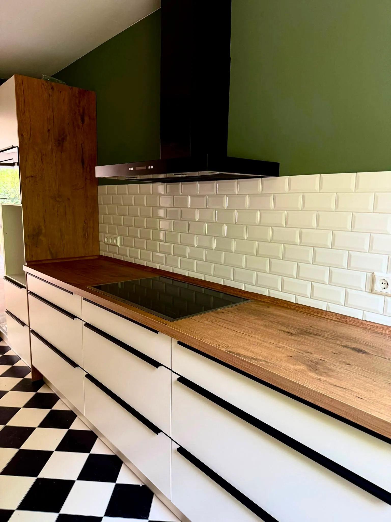 Modern kitchen with wooden countertops, white cabinets, black handles, a stovetop, and green walls with white subway tiles.