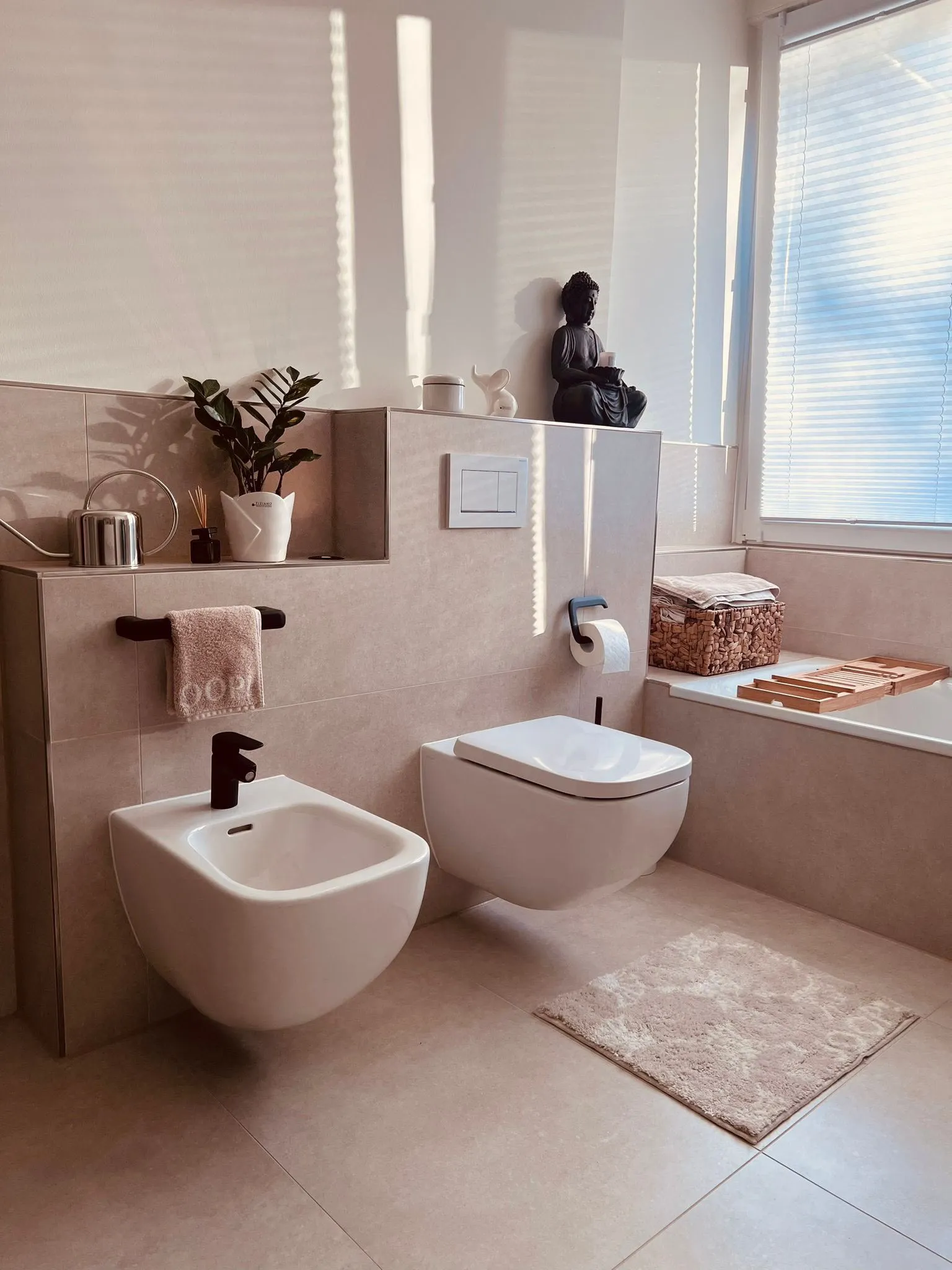 Modern bathroom with wall-mounted bidet and toilet, potted plant, Buddha statue, and natural light from window.