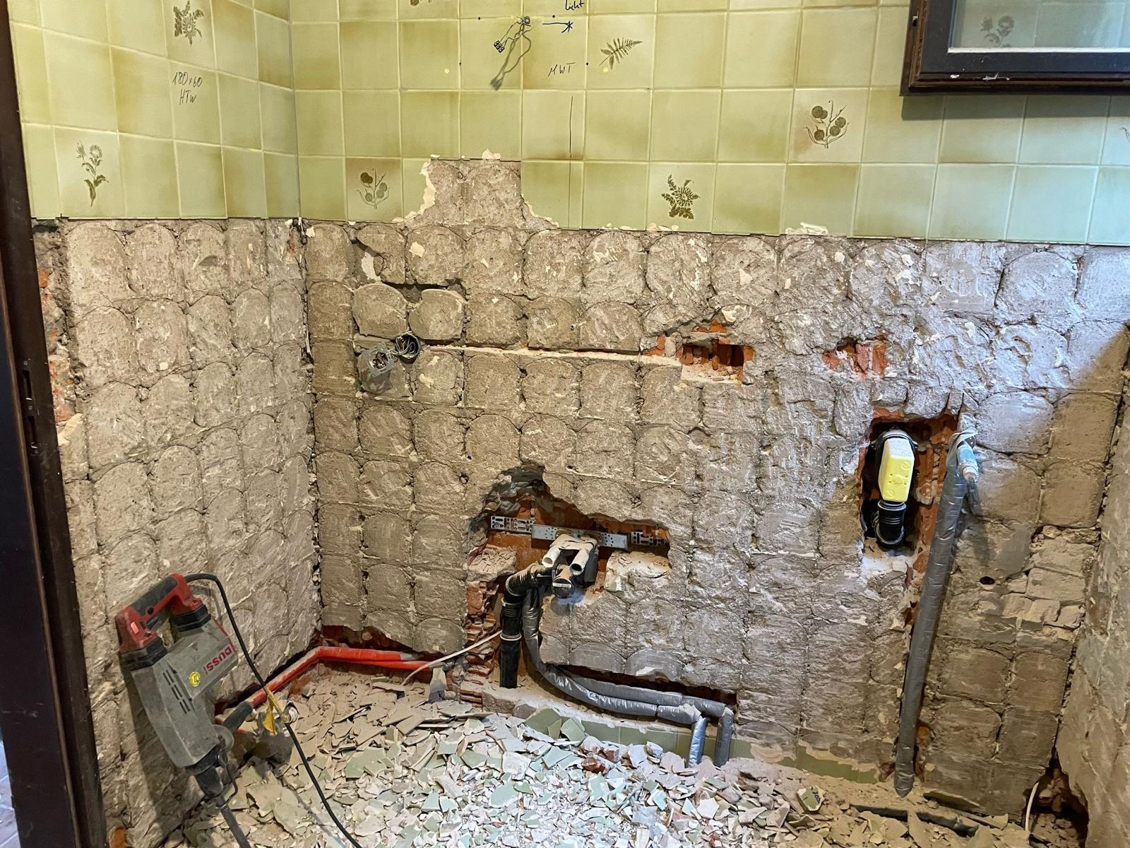 Room wall partially stripped of tiles showing old plaster, exposed pipes, electrical wires, and debris on floor.