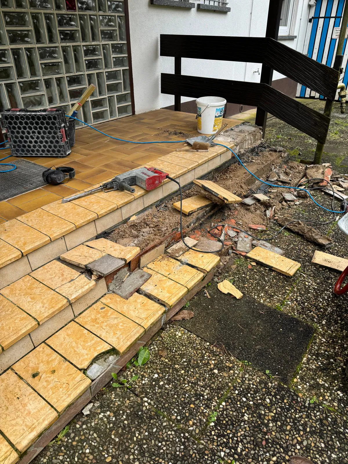 Broken outdoor tiled stairs under repair with tools, debris, and a bucket on a porch with glass bricks.