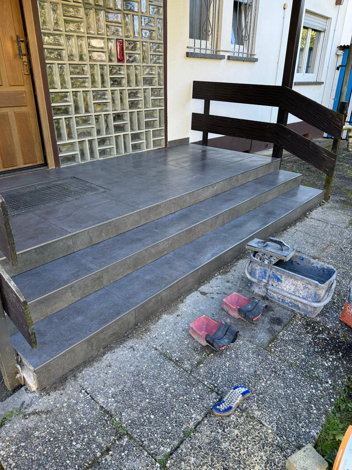Outdoor steps with gray tiles, wooden railings, glass block wall, and masonry tools on ground.