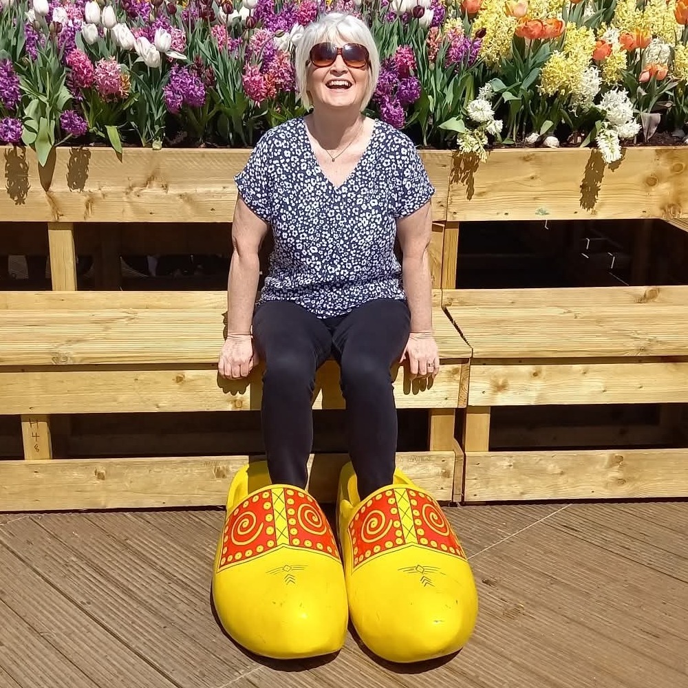 Linda laughing on a bench in giant yellow clogs, in front of a bed of tulips