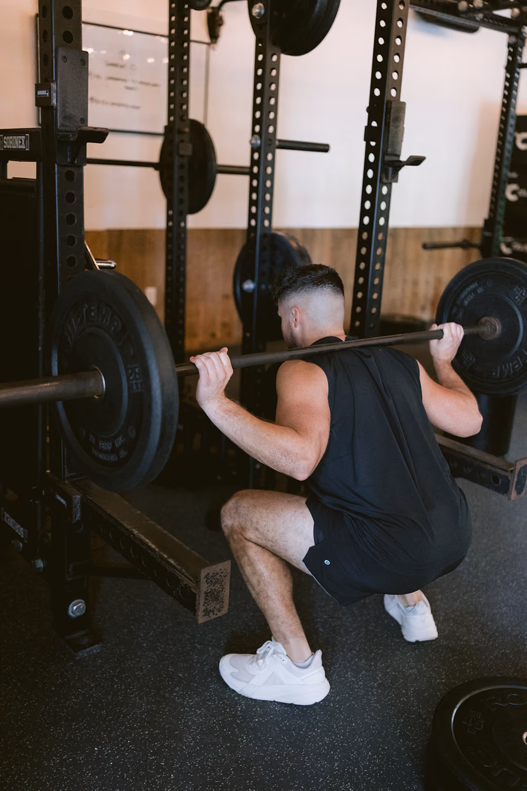 Man wearing black workout clothes and white sneakers performing a barbell squat in a gym with weight plates on the barbell and rack equipment in the background.