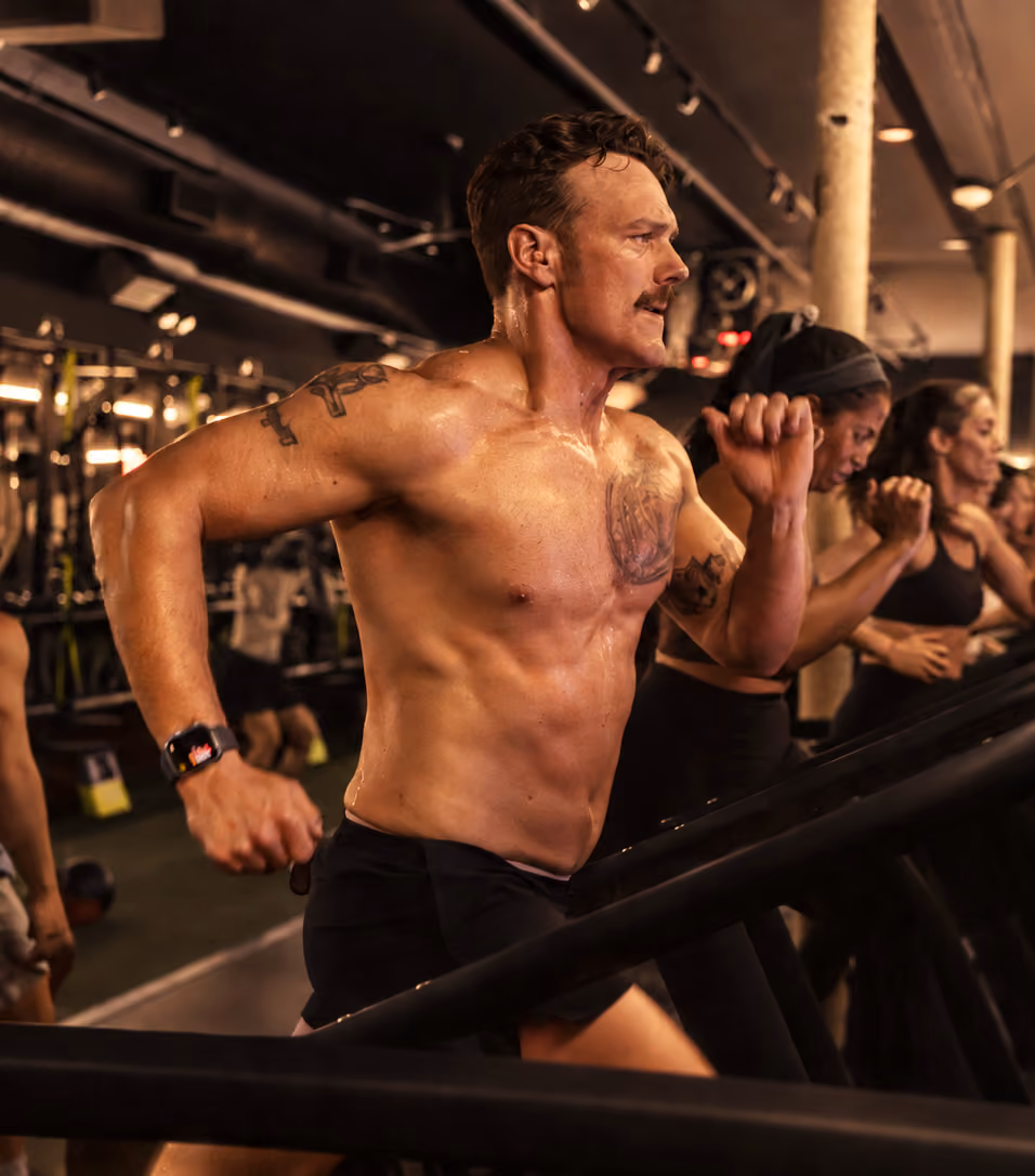 Shirtless muscular man with tattoos running intensely on a treadmill in a gym, with women exercising on treadmills beside him.