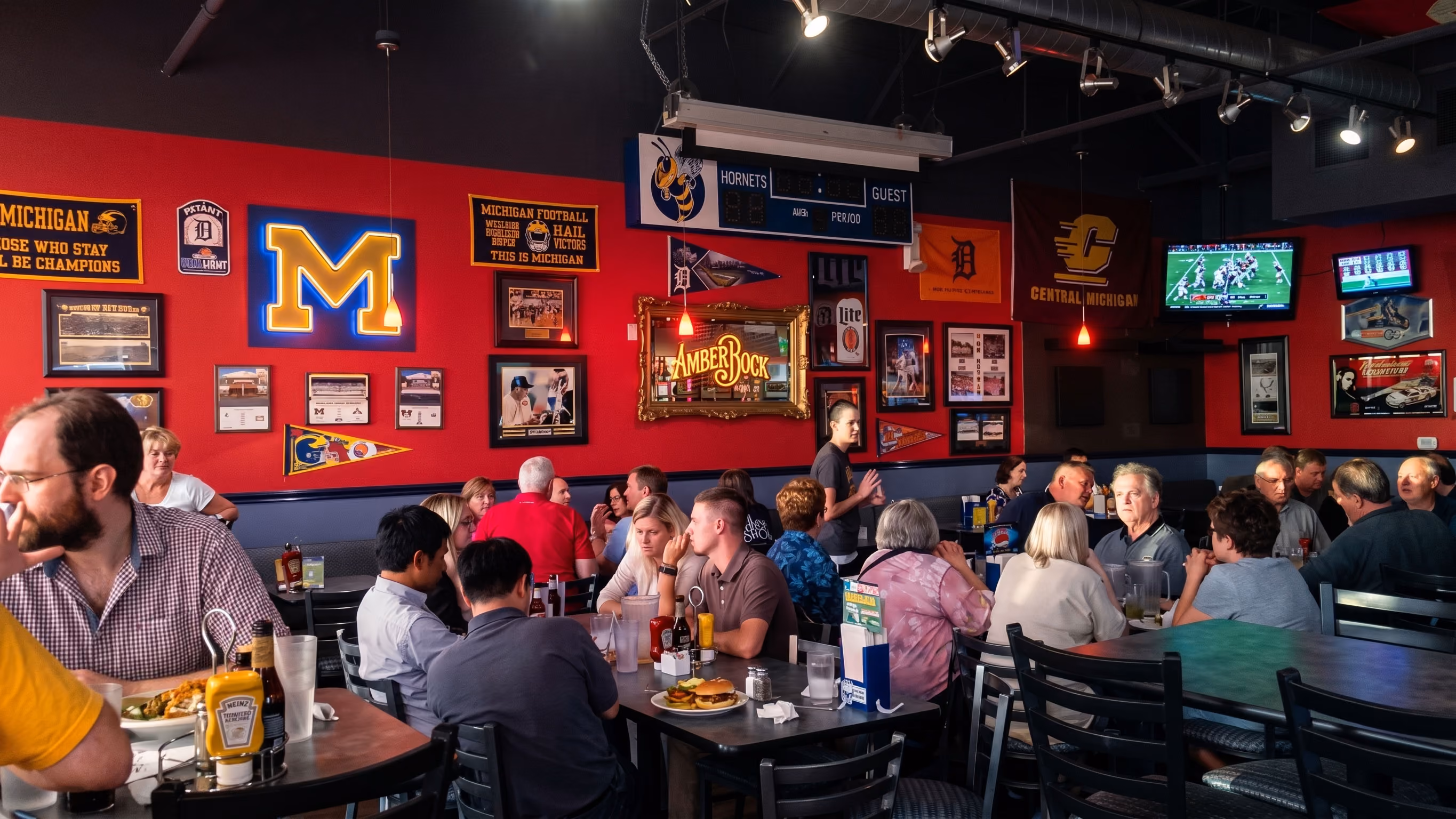 People dining in a sports-themed restaurant with red walls, sports flags, and TVs showing a football game.