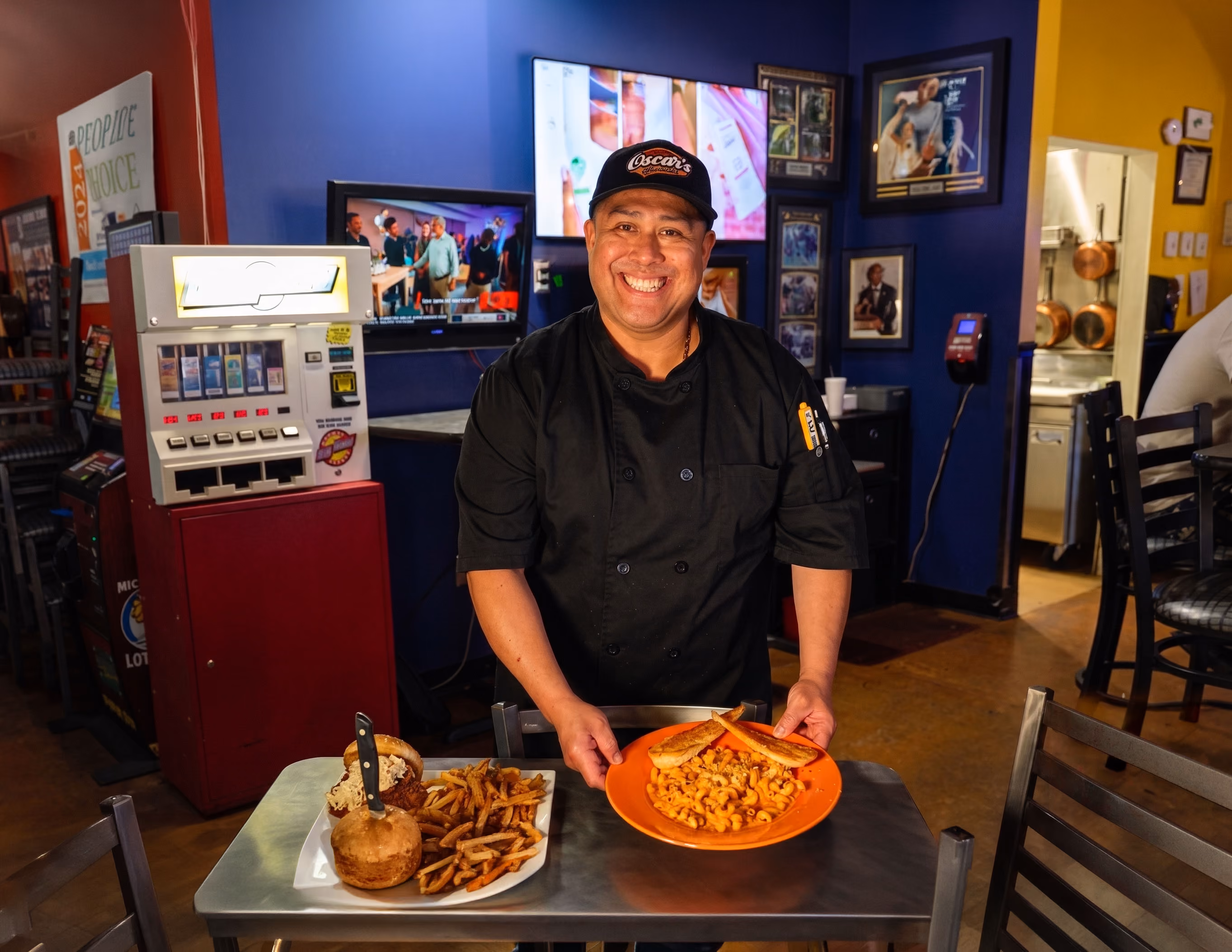 Smiling chef in black uniform holding a plate of elbow macaroni with bread in a colorful restaurant.