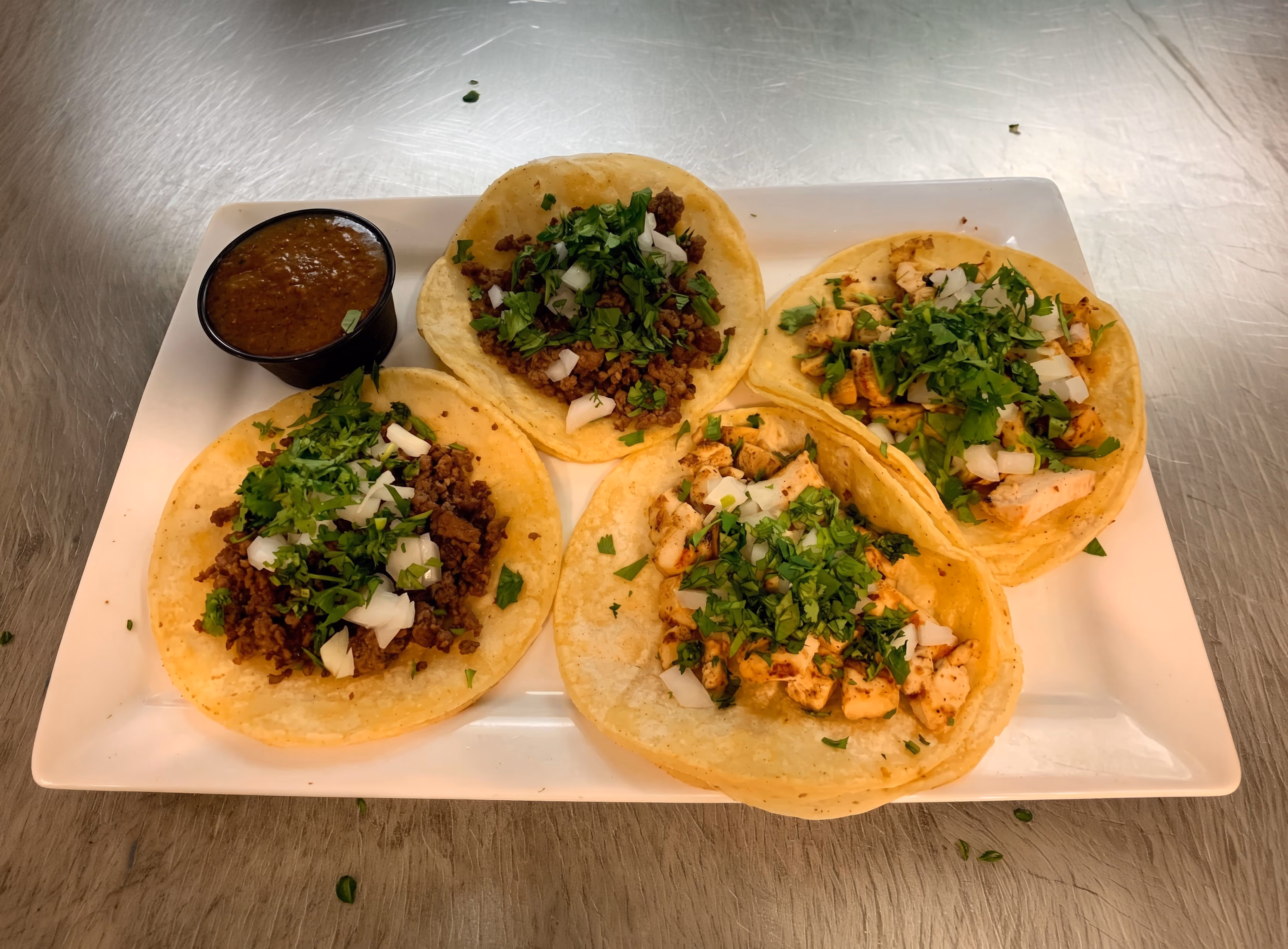 Plate with four street tacos topped with chopped onions and cilantro, served with a cup of salsa.