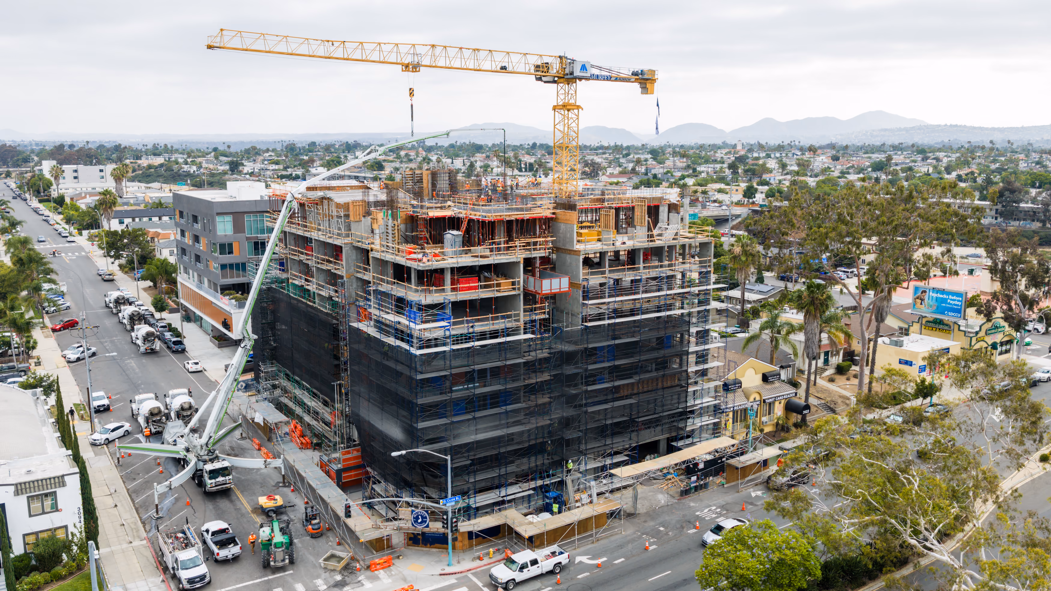 Large multi-story building under construction with cranes and workers in an urban neighborhood.