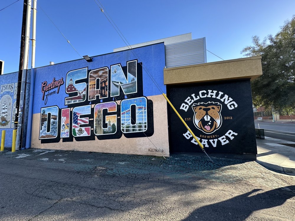 Colorful mural saying Greetings from San Diego with images inside letters next to Belching Beaver Brewery logo.