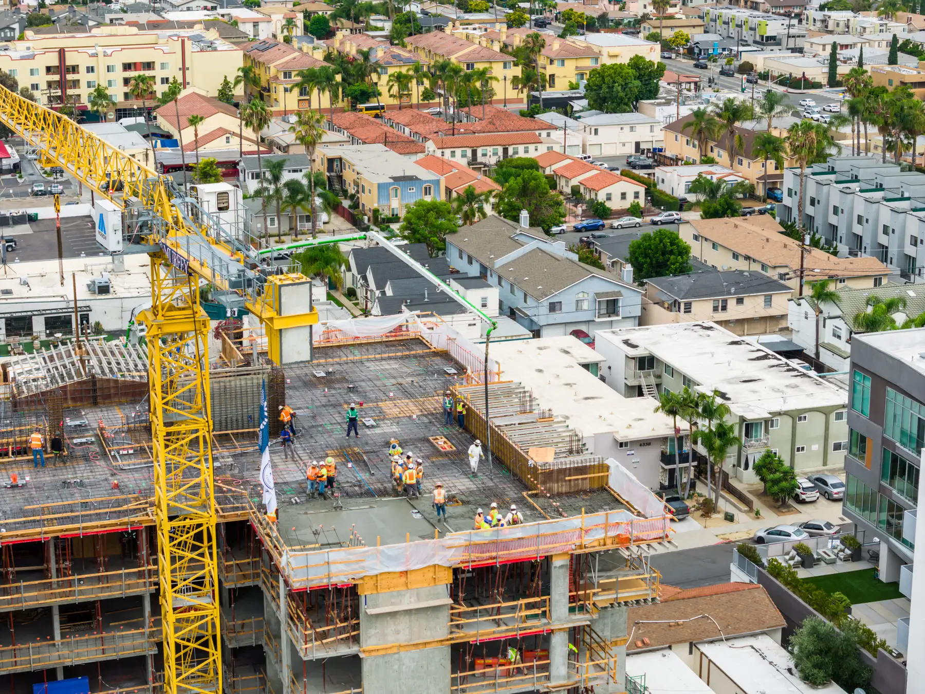 Construction workers on a high-rise building floor with a yellow crane in a dense residential neighborhood.