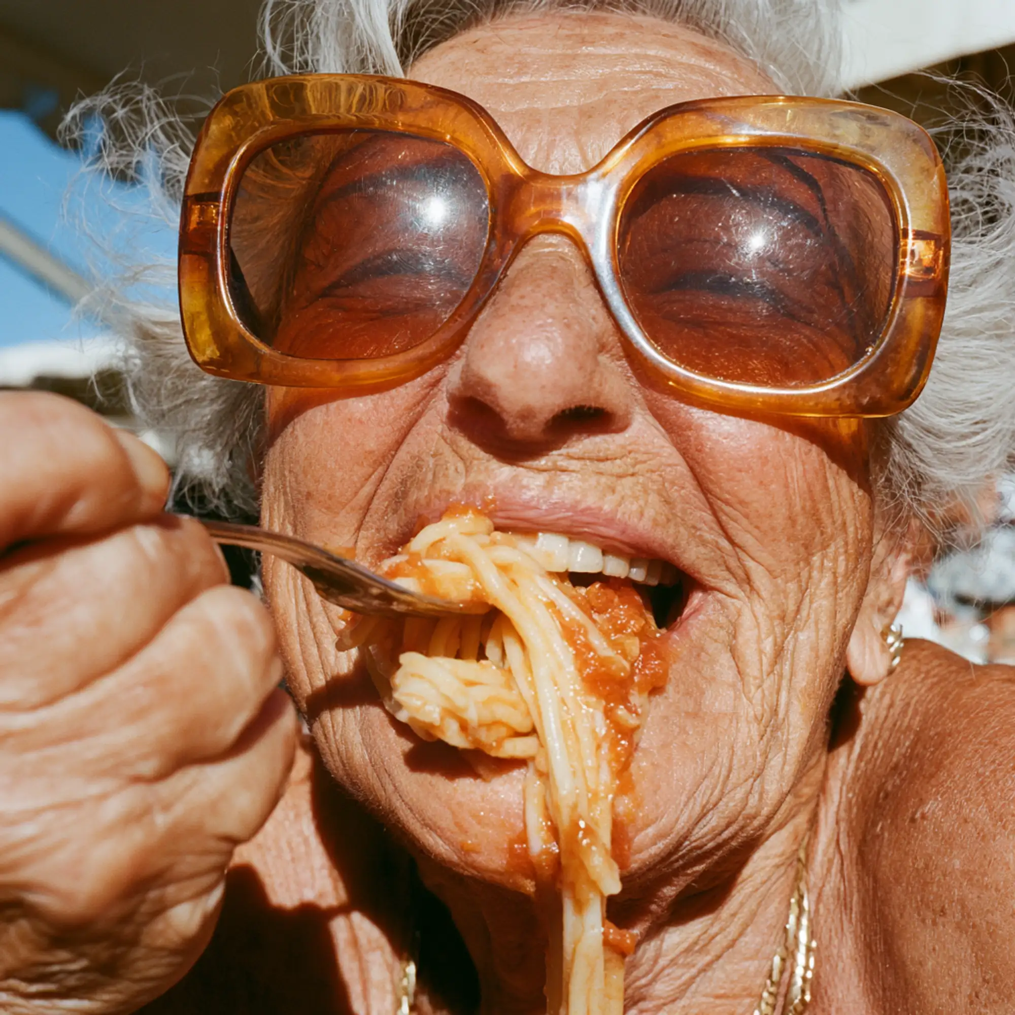 Elderly woman wearing large amber sunglasses eating spaghetti with tomato sauce outdoors.