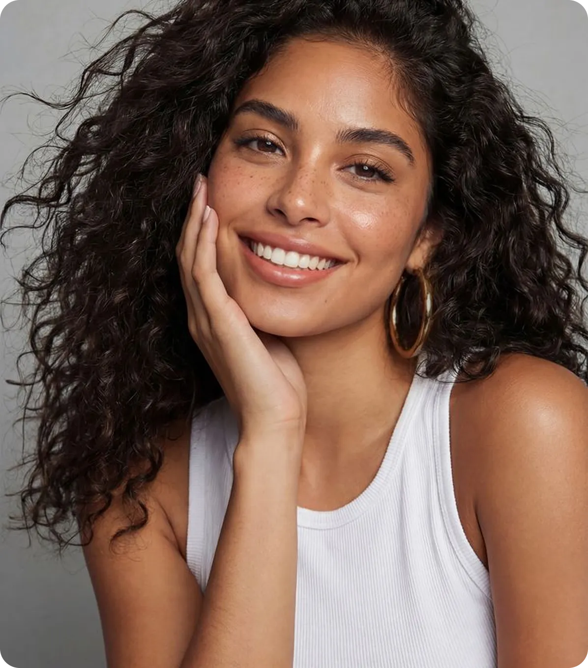 Smiling woman with curly dark hair, freckles, gold hoop earrings, and a white sleeveless top.