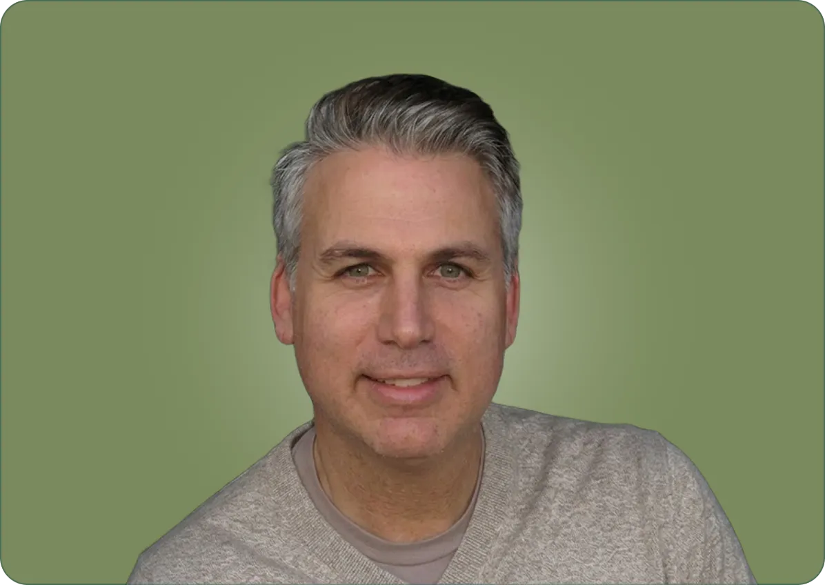 Smiling middle-aged man with gray hair wearing a beige sweater against a muted green background.