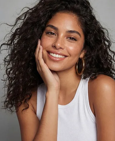 Smiling woman with curly hair wearing hoop earrings and a white sleeveless top against gray background.