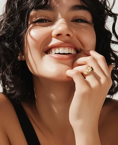 Close-up of a smiling woman with curly hair, wearing a gold ring and a black strap top.