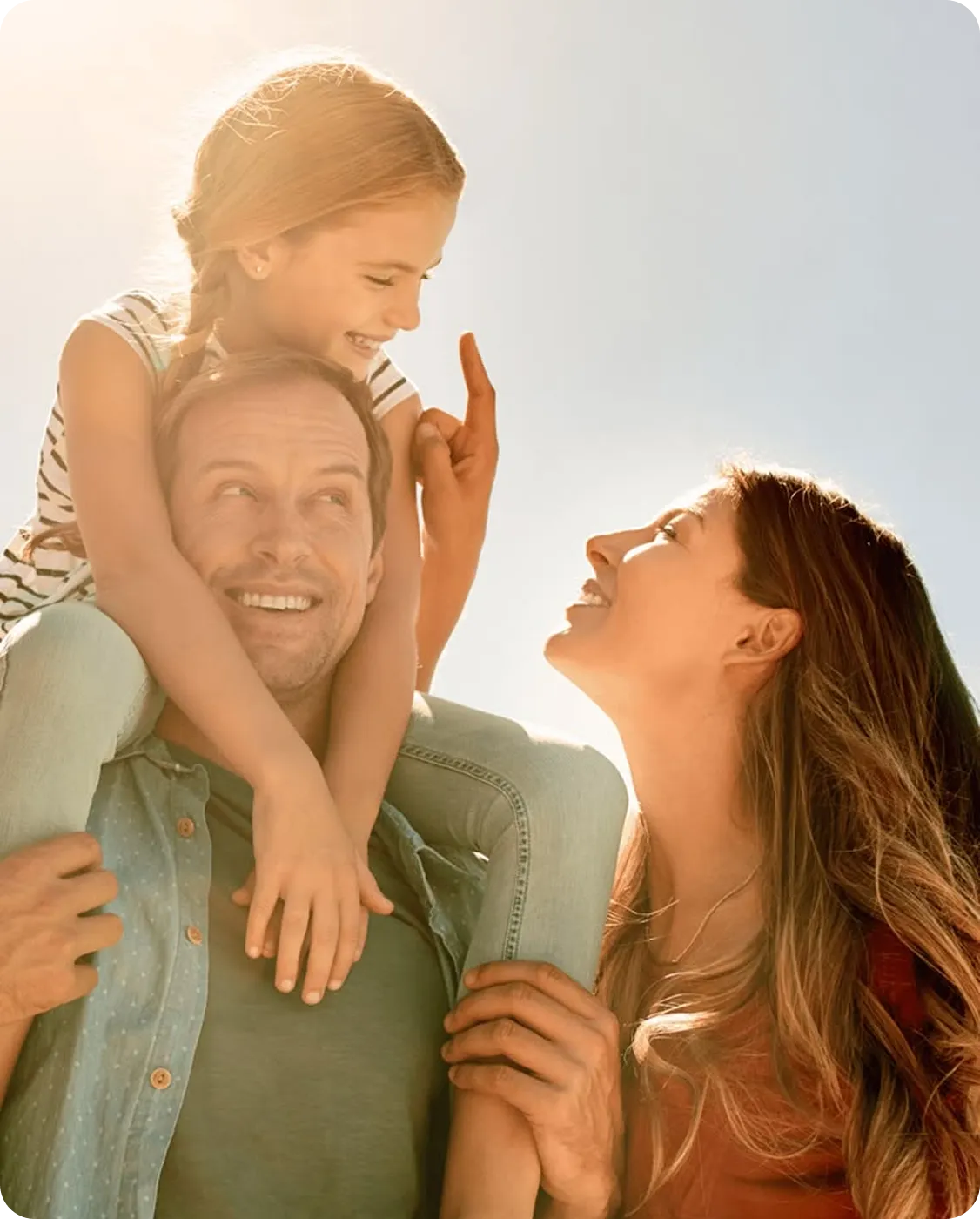 Smiling little girl on man's shoulders holding his head, woman smiling and looking up at them outdoors.